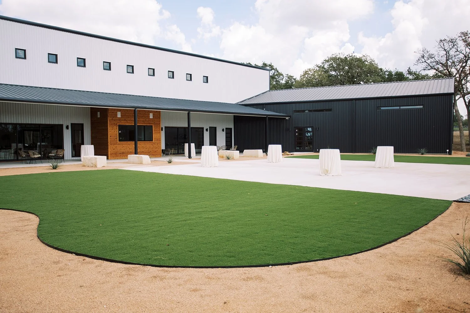 Modern building with black and white exterior, large patio area with tall tables covered in white cloths, and shaded seating area. The yard has manicured green turf and beige sandy pathways.