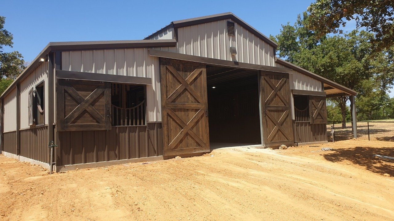A large barn with sliding wooden doors, metal siding in beige and brown, and a dirt driveway in front. The barn has a covered outdoor area on the right side and is surrounded by trees on a sunny day.