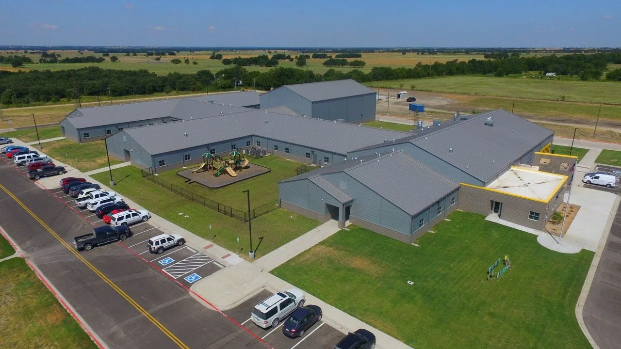 Aerial view of a school building with a playground, parking lot, and surrounding green fields.
