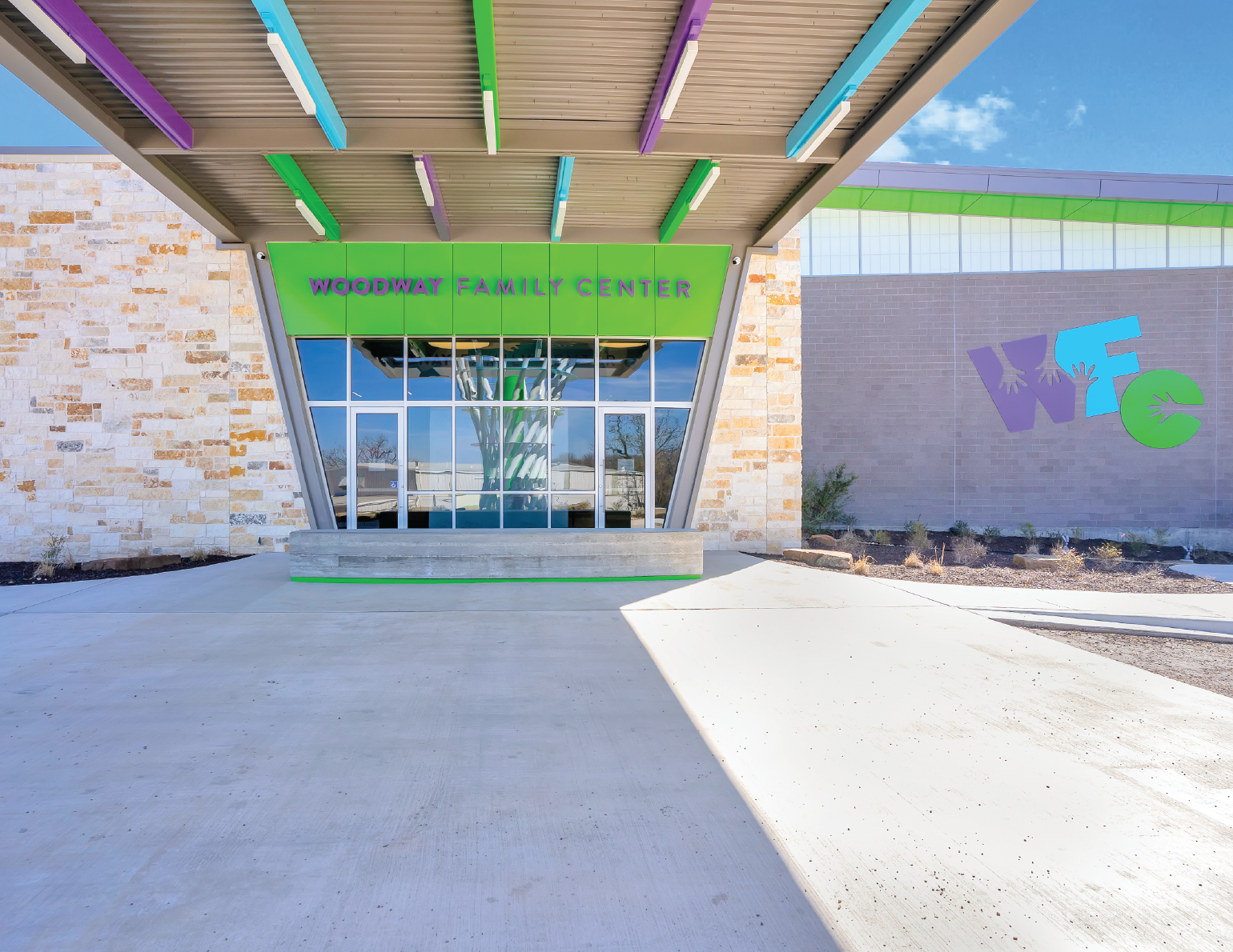 Exterior of Woodway Family Center with colorful signage and modern architectural design, including large glass windows, stone accents, and a mural with the letters WEC.