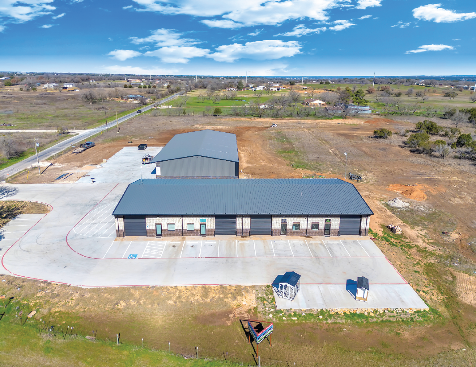 An aerial view of a newly constructed commercial or industrial building with a blue metal roof, paved parking lot, and surrounding landscape, with open fields and a partly cloudy sky in the background.