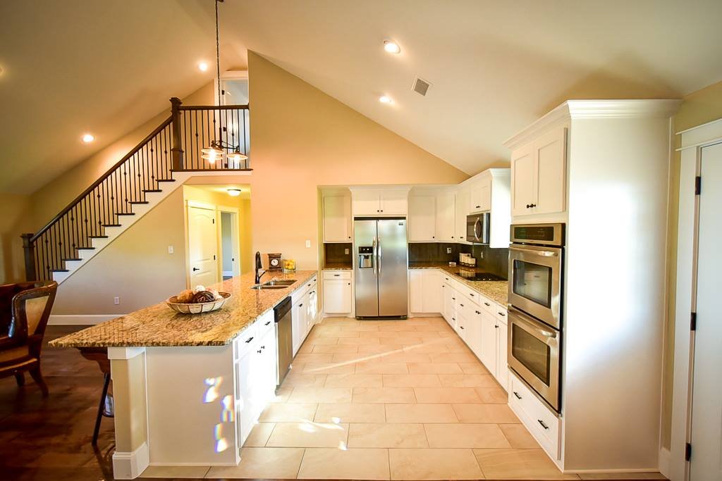Kitchen with white cabinets, granite countertops, stainless steel appliances, and a tiled floor. An island with a fruit basket and a seating area is in the foreground. A staircase with dark wooden railing leads upstairs.