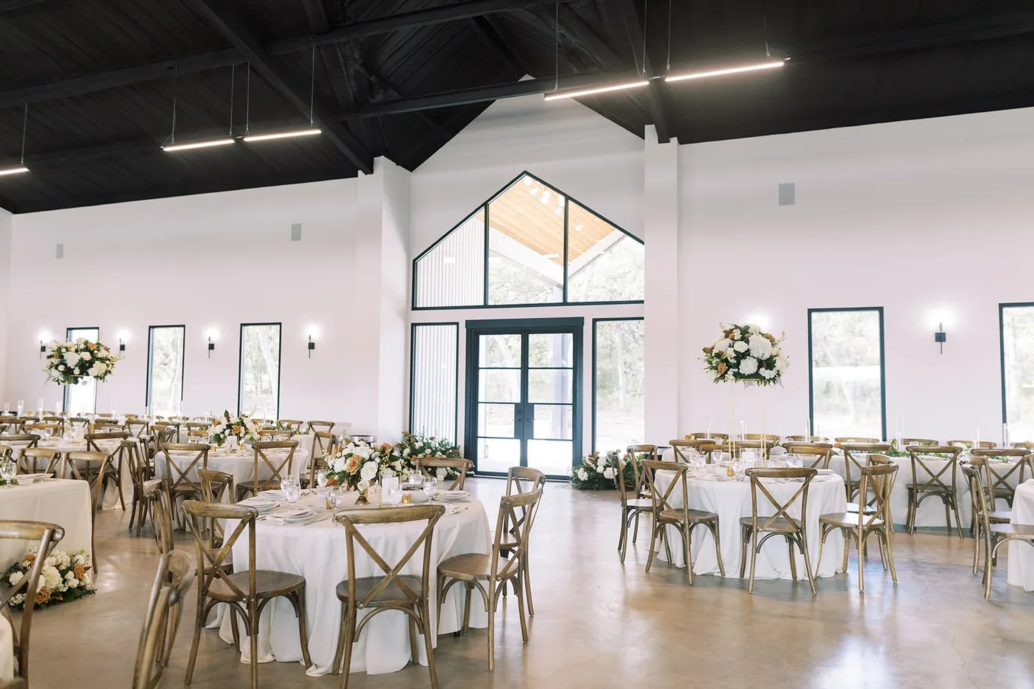 Elegant event space with round tables decorated with white tablecloths, floral centerpieces, and wooden chairs. The room has large windows, white walls, and high black ceiling with modern lighting fixtures.