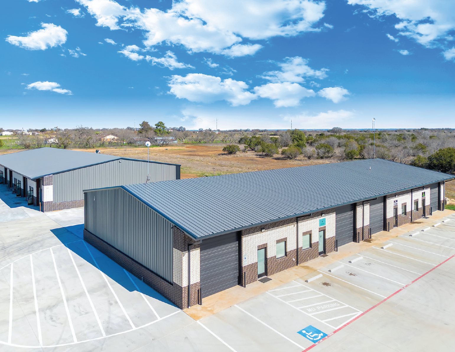 An aerial view of a modern commercial building with a metal roof and brick walls, surrounded by parking spaces, including a handicap spot, under a partly cloudy sky.