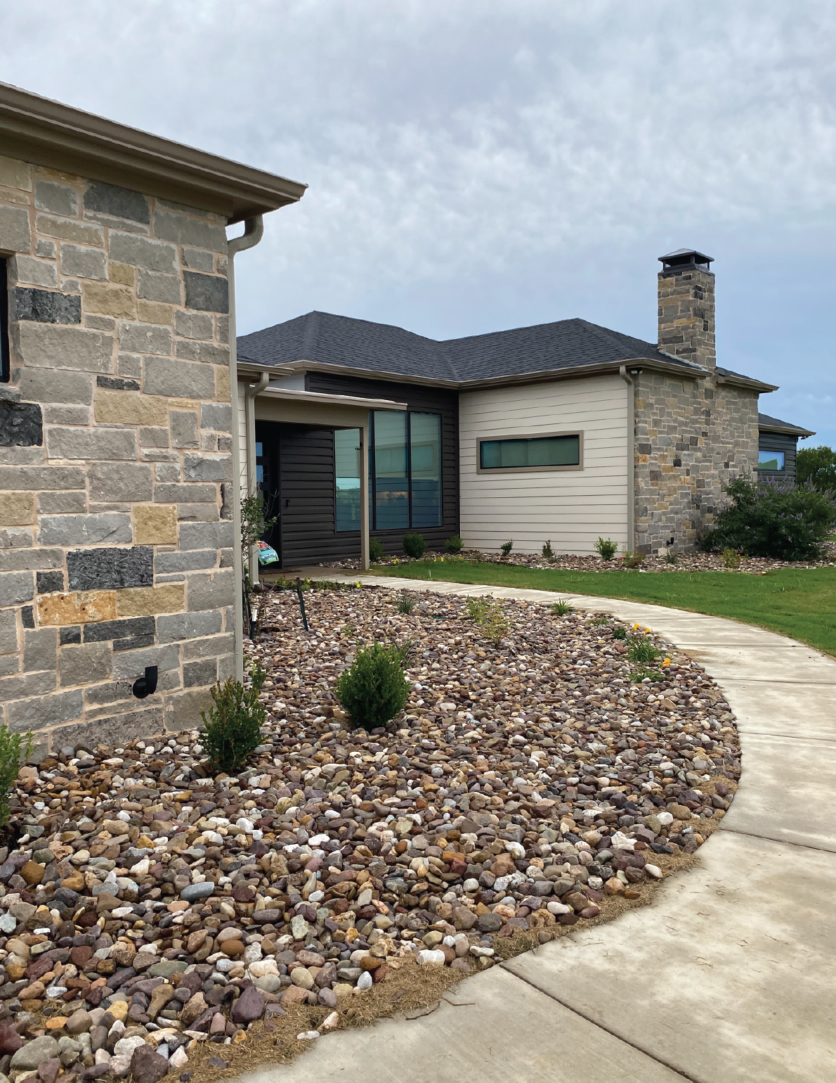 View of modern house with stone and siding exterior, landscaped yard with gravel, small shrubs, and a curved concrete walkway.