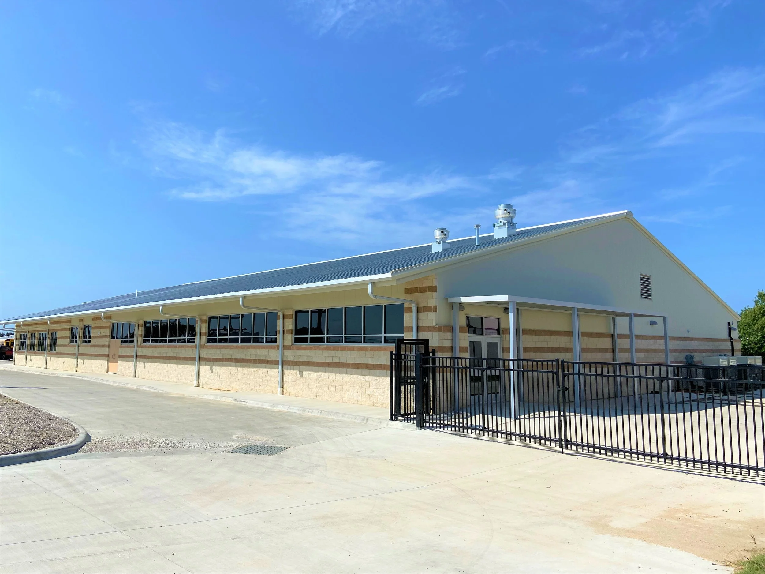 A single-story building with a brick lower wall and a light blue upper wall, featuring large windows and a sloped metal roof, surrounded by a black metal fence under a clear blue sky.