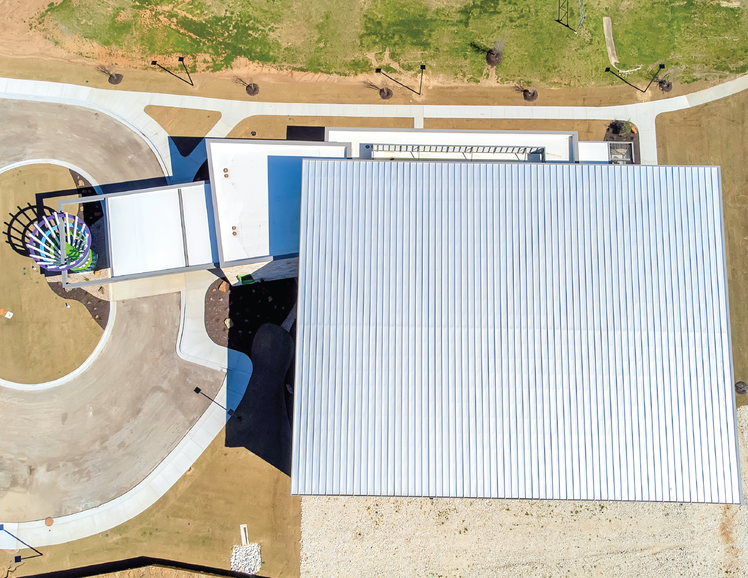 Aerial view of a modern building with a metal roof, a playground with a climbing structure, and surrounding pathways with trees and grass.