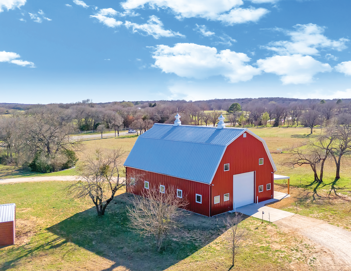 An aerial view of a red barn with a blue metal roof, surrounded by a rural landscape with leafless trees, open fields, and a clear blue sky.