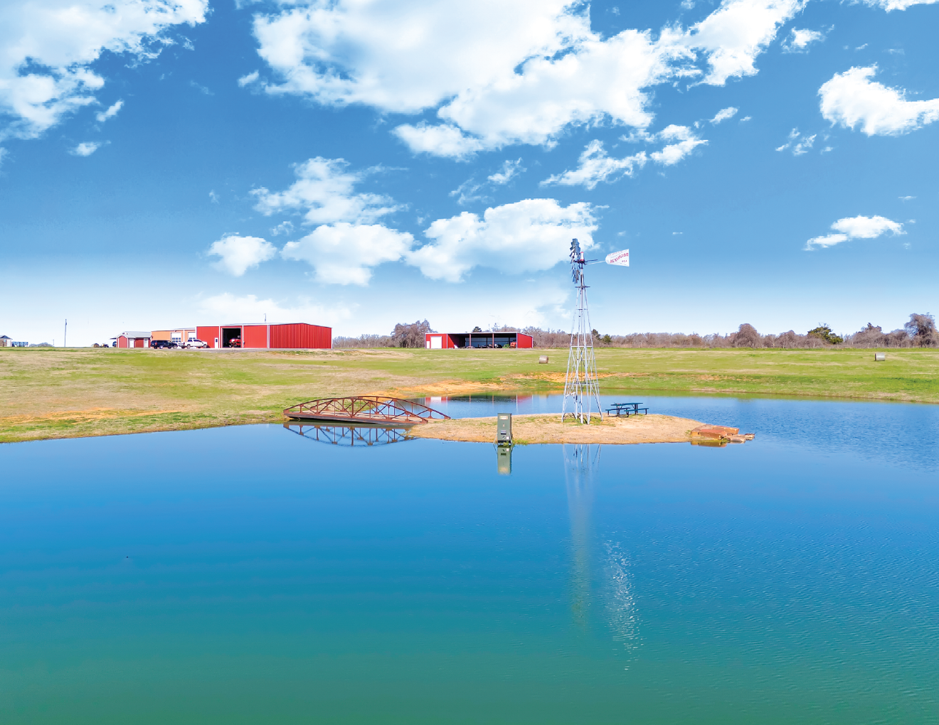 A rural landscape with a small pond, a windmill, a bridge, and red barn buildings in the background under a partly cloudy sky.