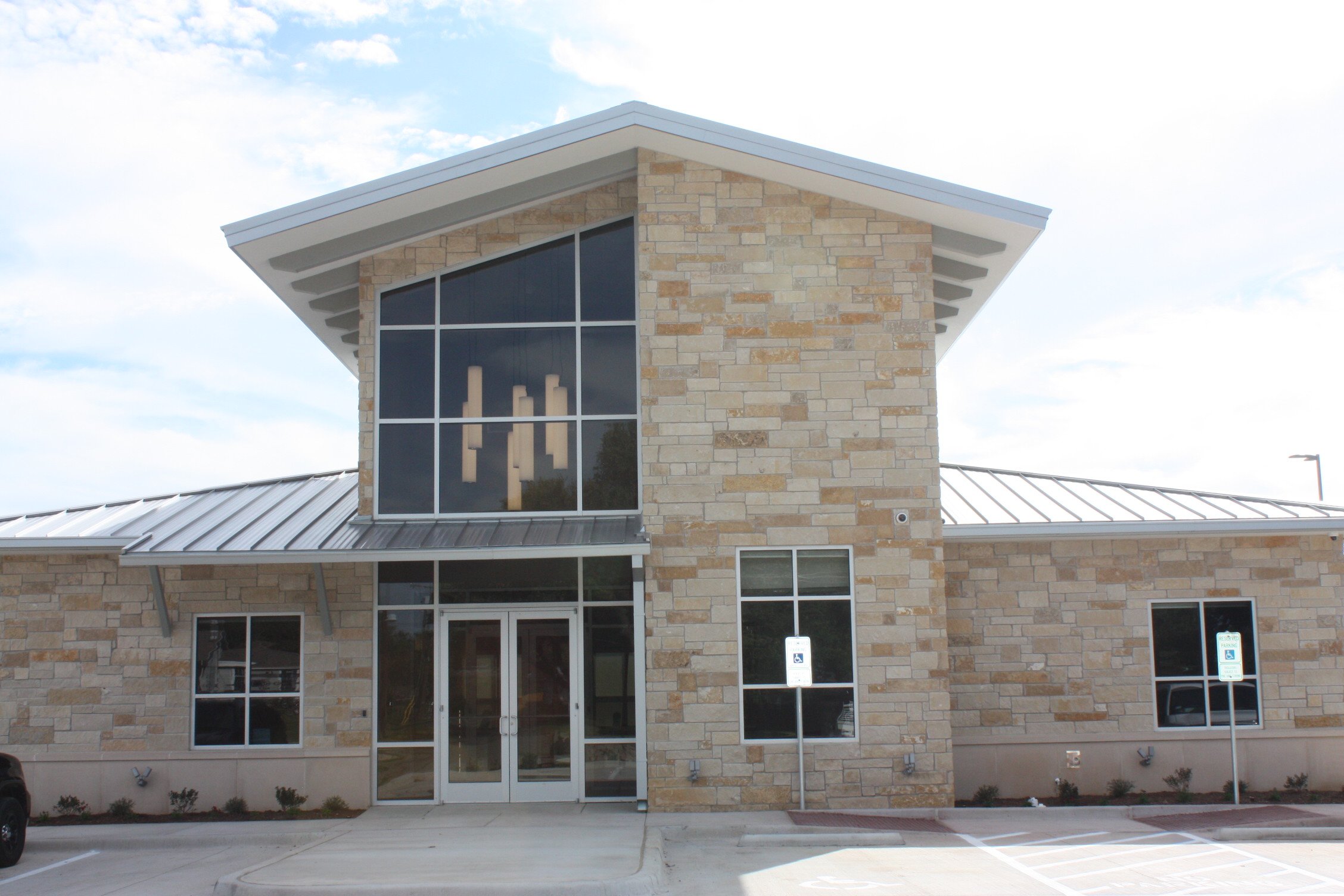 Modern building with stone exterior, large glass windows, and a metal roof, with accessible parking spaces in front.
