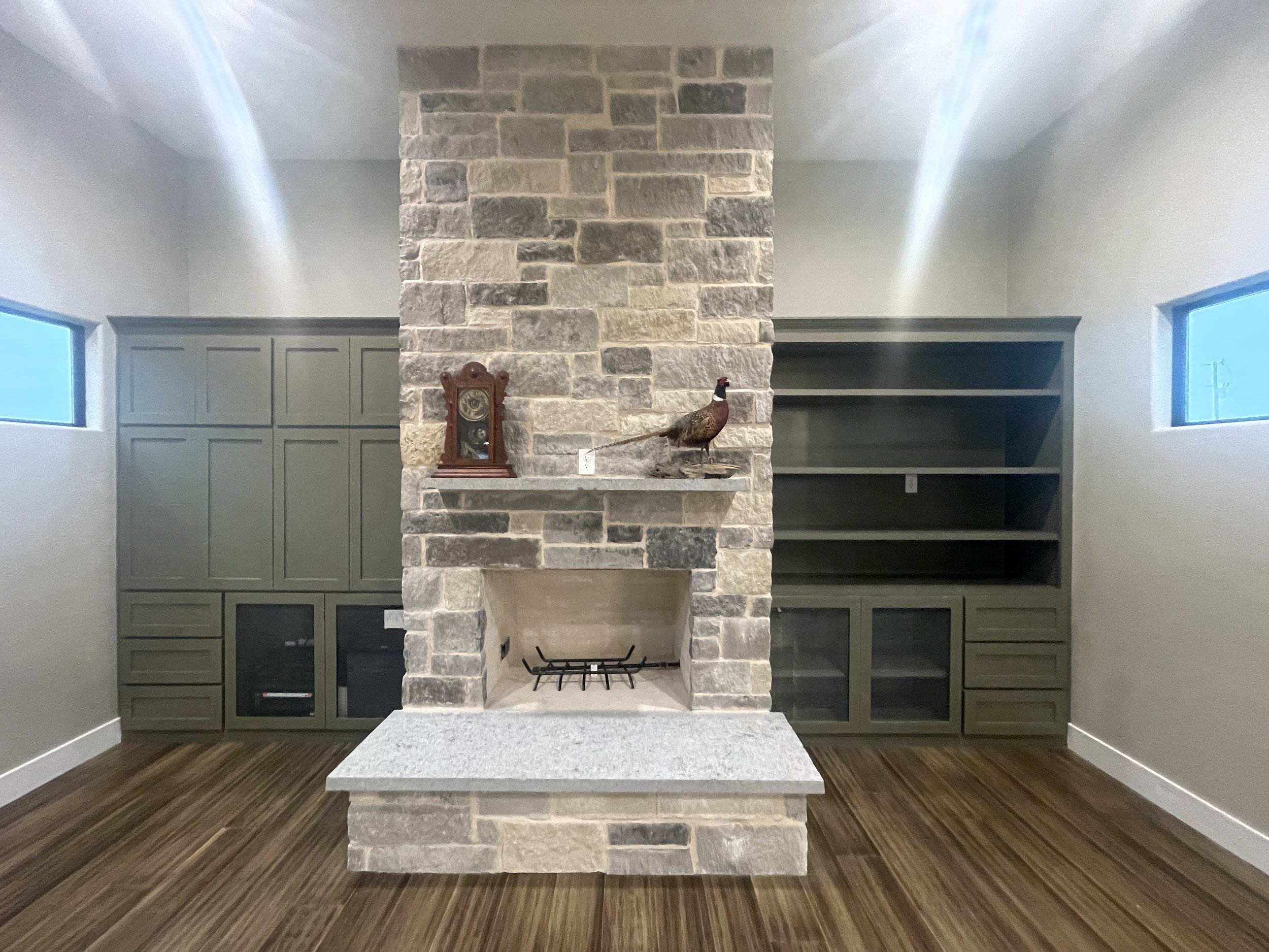 Living room with stone fireplace, built-in shelves and cabinets, small windows, and wooden flooring.