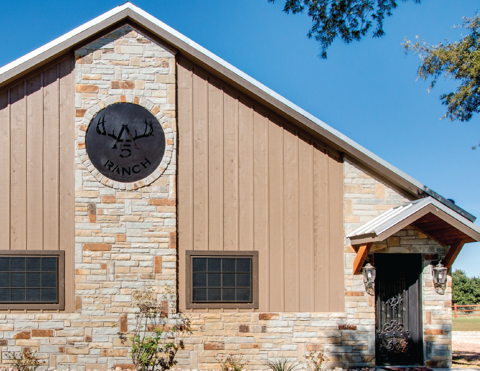 A ranch building with a stone and wood facade, featuring a large round clock with antlers and the words '5 Ranch', two small windows, and an ornate black gate entrance under a small gabled roof, against a clear blue sky.