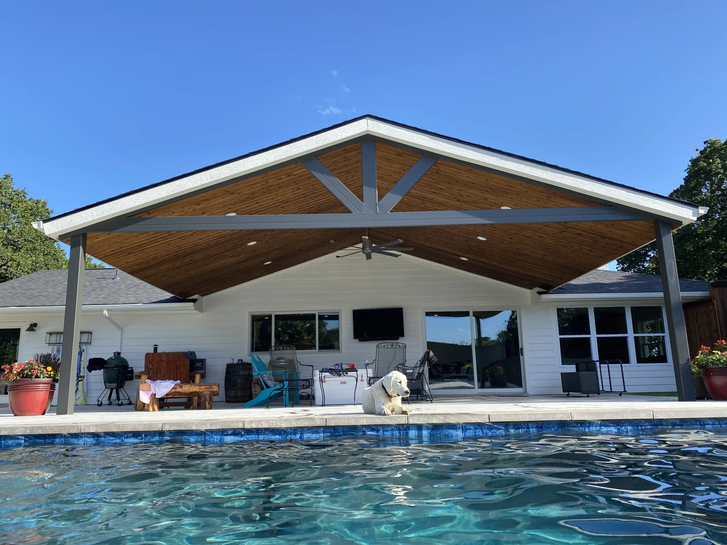Backyard with swimming pool, covered patio, outdoor furniture, potted plants, a TV, and a dog laying near the pool, under a clear blue sky.