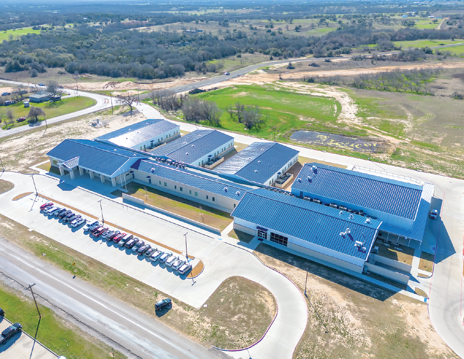Aerial view of a new school complex with multiple buildings, parking lot, and surrounding undeveloped land.