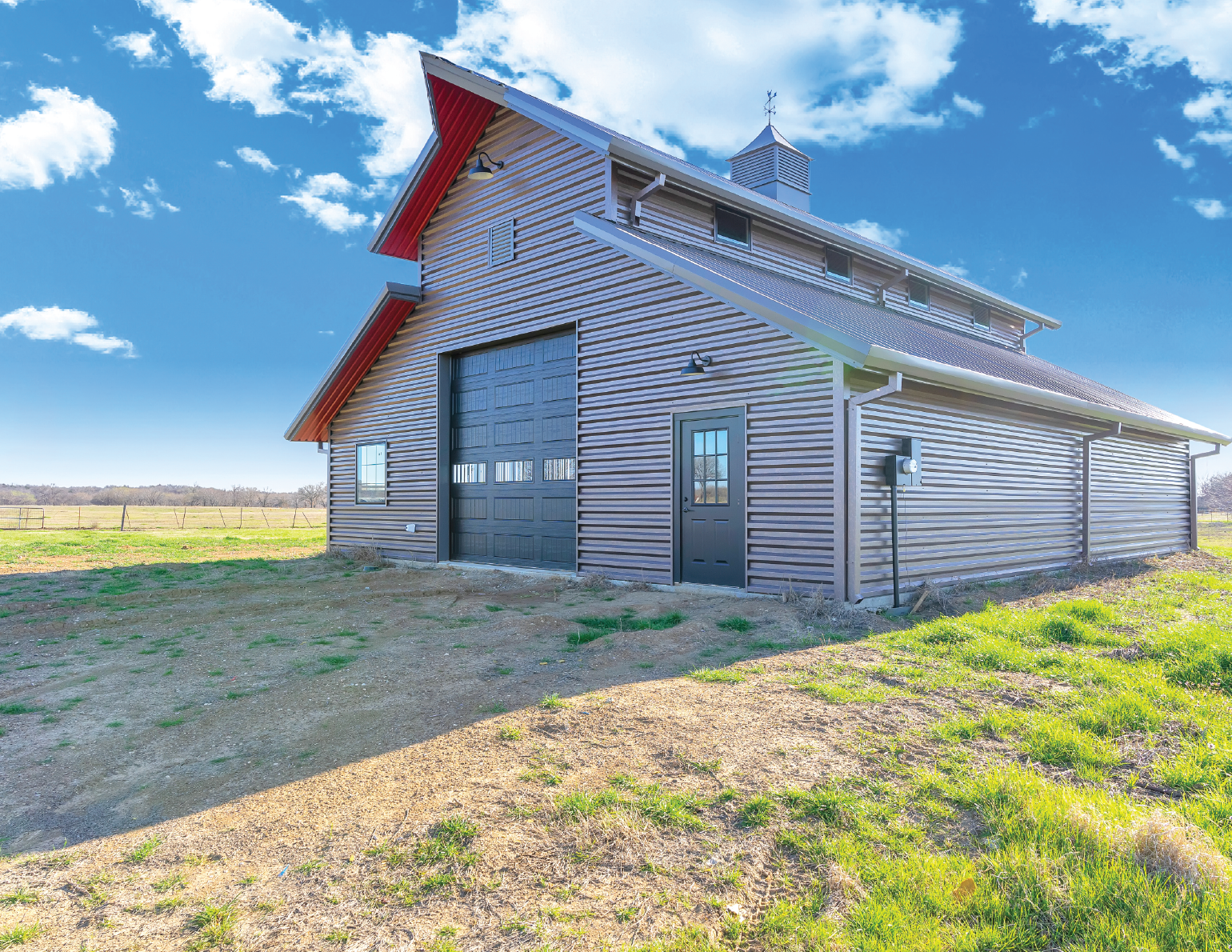 A large barn with a black garage door, smaller door, and windows, situated in an open field under a partly cloudy blue sky.