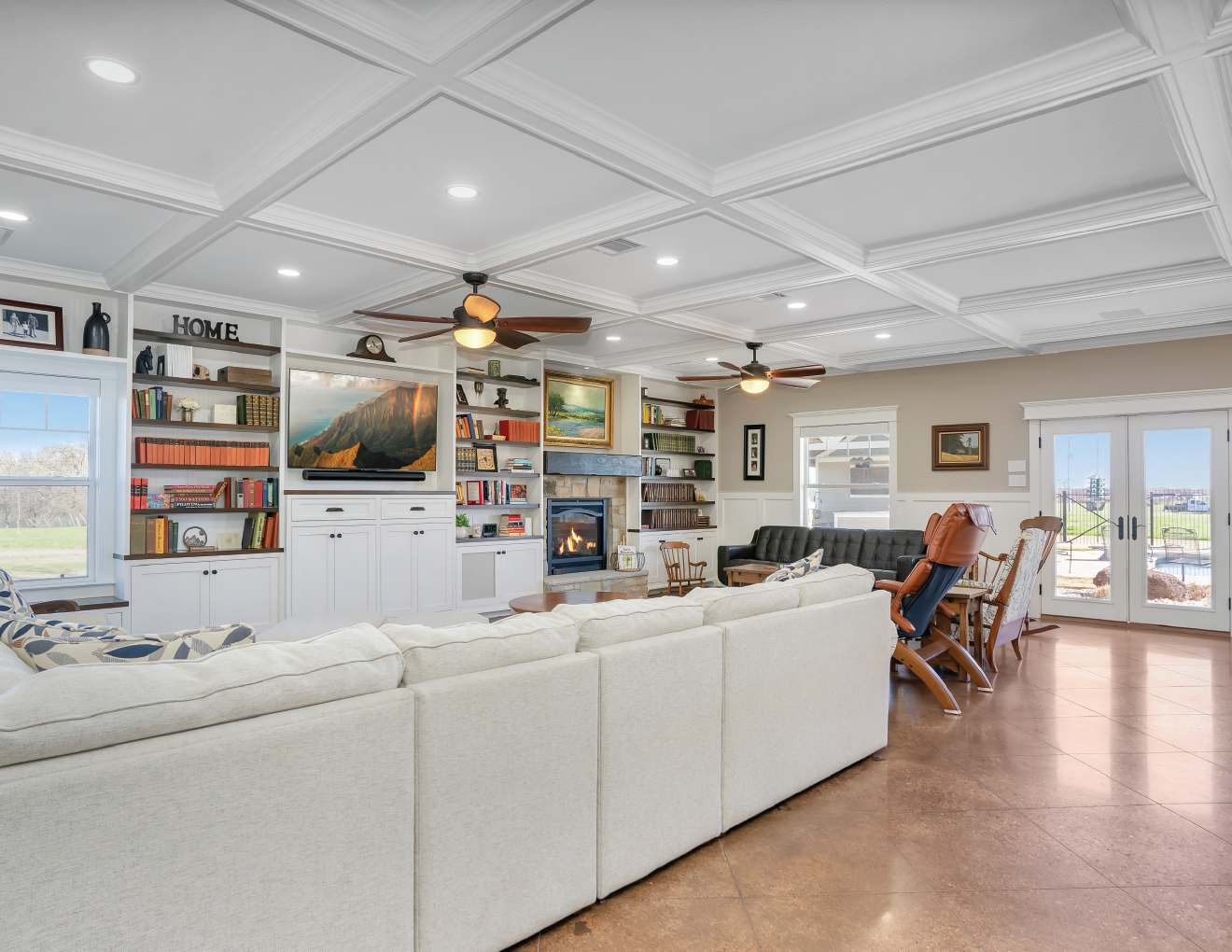 Living room with white sectional sofa, built-in white bookshelves, a fireplace, a mounted TV, and multiple ceiling fans with lights. Large windows and glass doors offer a view of a green outdoor area.