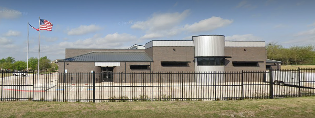 Exterior view of a modern building with brick walls, a cylindrical metal section, and a black fence in front, with an American flag on a flagpole.