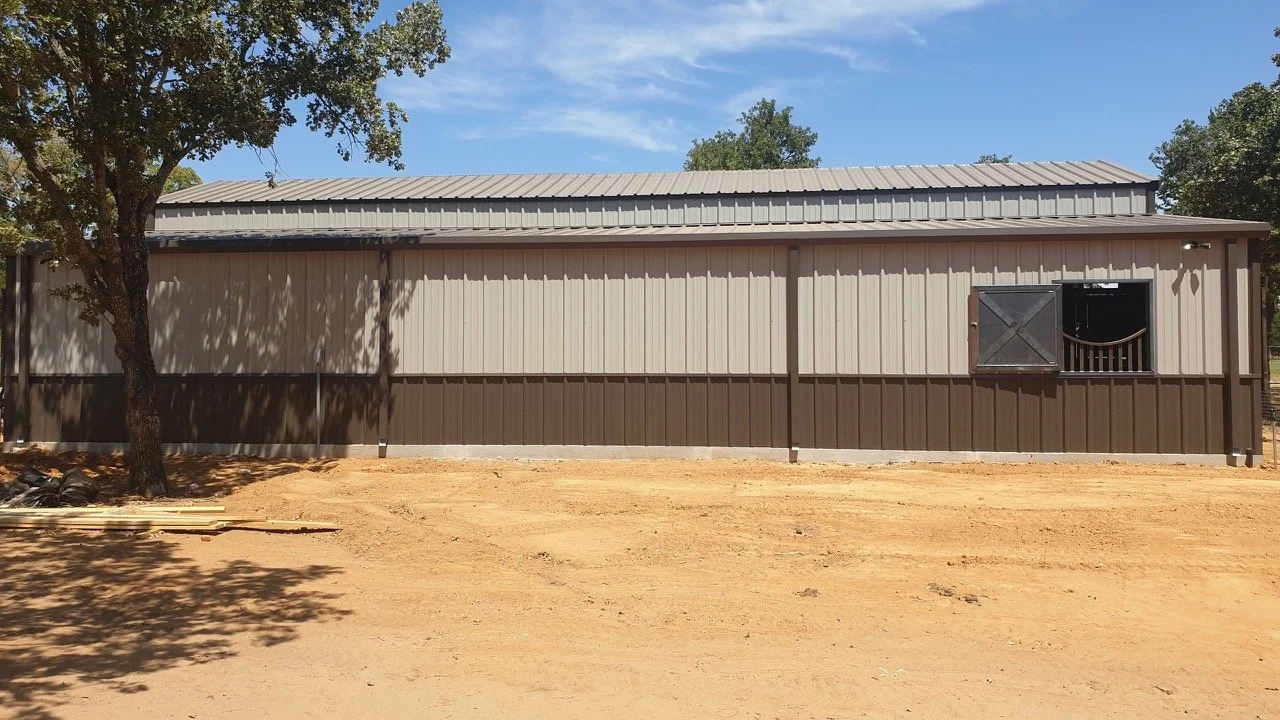 A beige and brown metal shed or barn with an open window and a tree on the left side, on a dirt ground under a blue sky with some clouds.