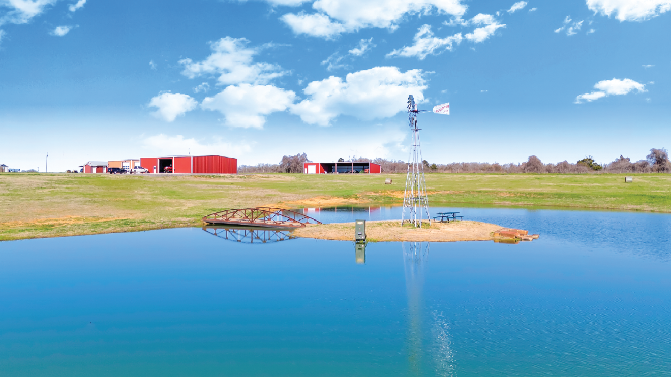 A pond with a small wooden bridge, a windmill, and a picnic table in the foreground, with red barn buildings and a blue sky with clouds in the background.
