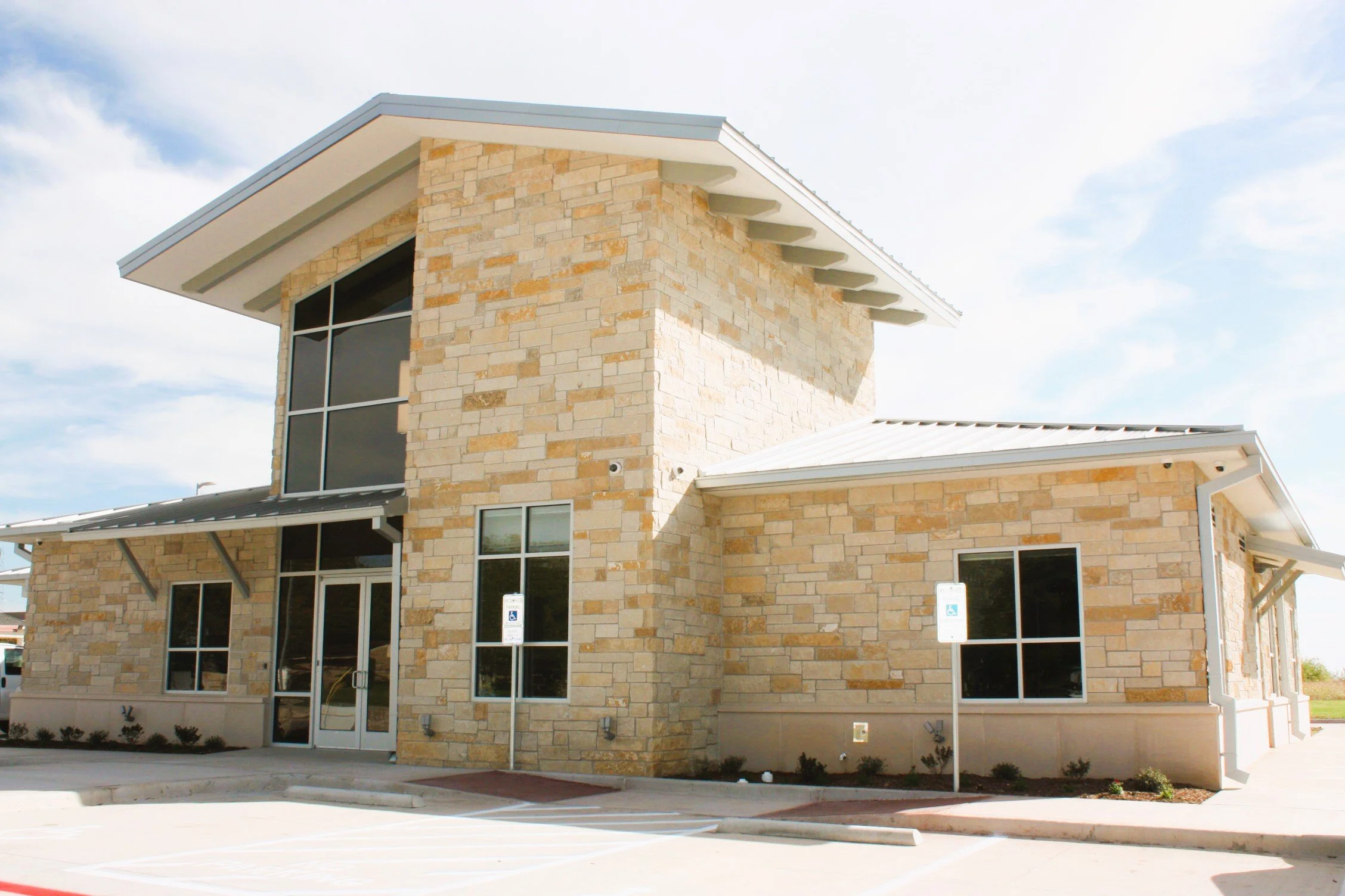 Modern two-story building with a stone exterior, large windows, and a metal roof, set against a partly cloudy sky.