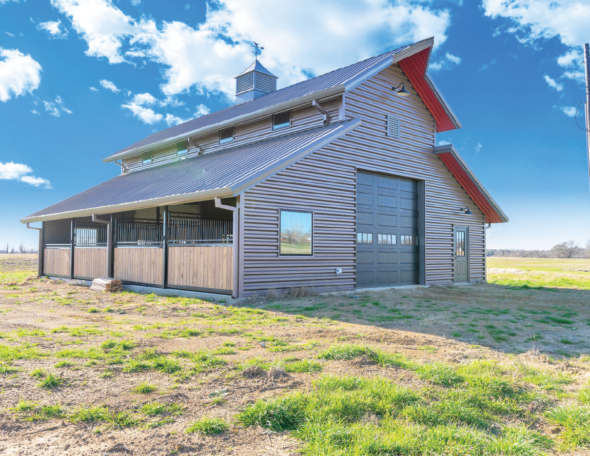 A large barn with metal and wood siding, black garage door, small windows, and a metal cupola on a rural field under a partly cloudy sky.