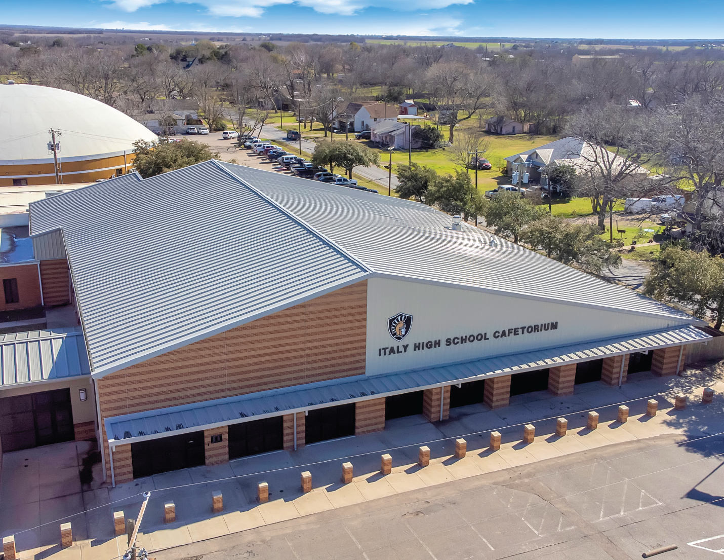 Aerial view of Italy High School Cafetorium with a metal roof, beige brick walls, and a sign displaying the school's name and emblem, surrounded by a parking lot.