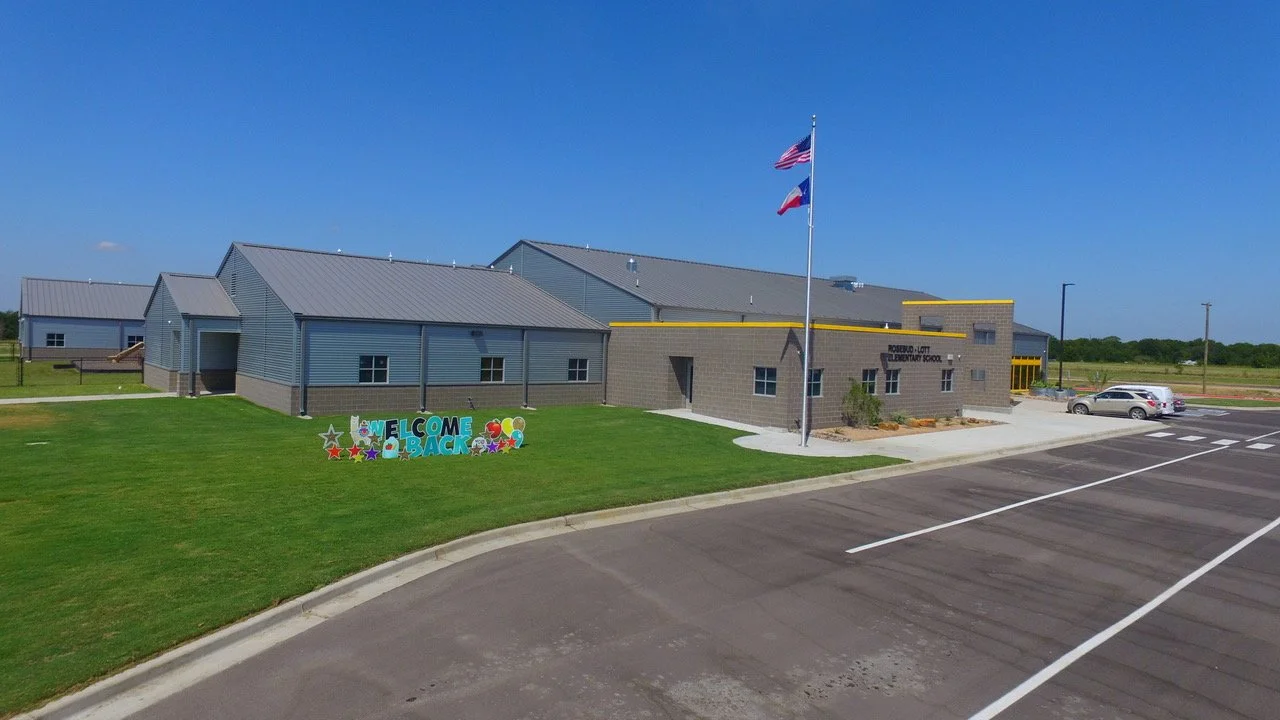Newly built Elementary School with a sign that says "Welcome Back" on the lawn, flags of the United States and Texas, and a parking lot with a few cars under a clear blue sky.