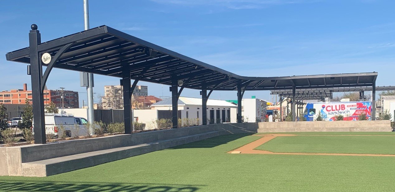 A baseball field with a protective covered seating area, artificial turf, and a paved base path. In the background, there are buildings, a truck, and a sign for a sandwich shop.