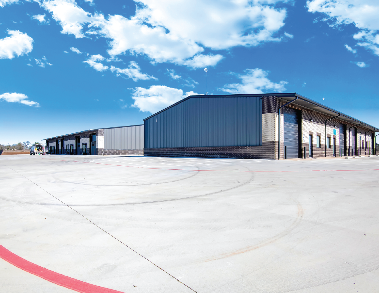 Large warehouse building with metal siding and brick accents, set against a blue sky with scattered clouds, and a wide concrete parking lot in the foreground.