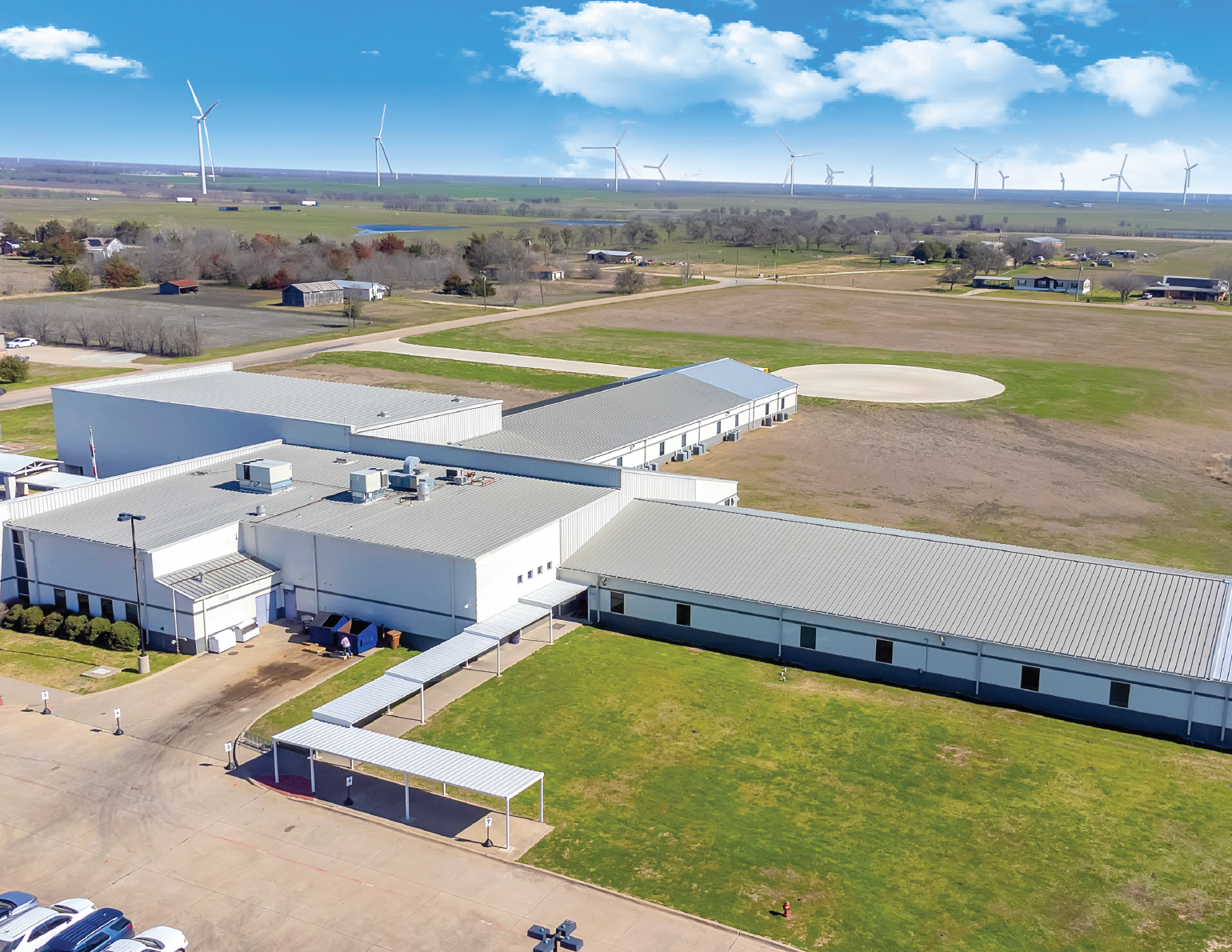 Aerial view of a large white industrial building with metal roofs, parking lot, and a grassy area, with wind turbines on the horizon under a partly cloudy sky.