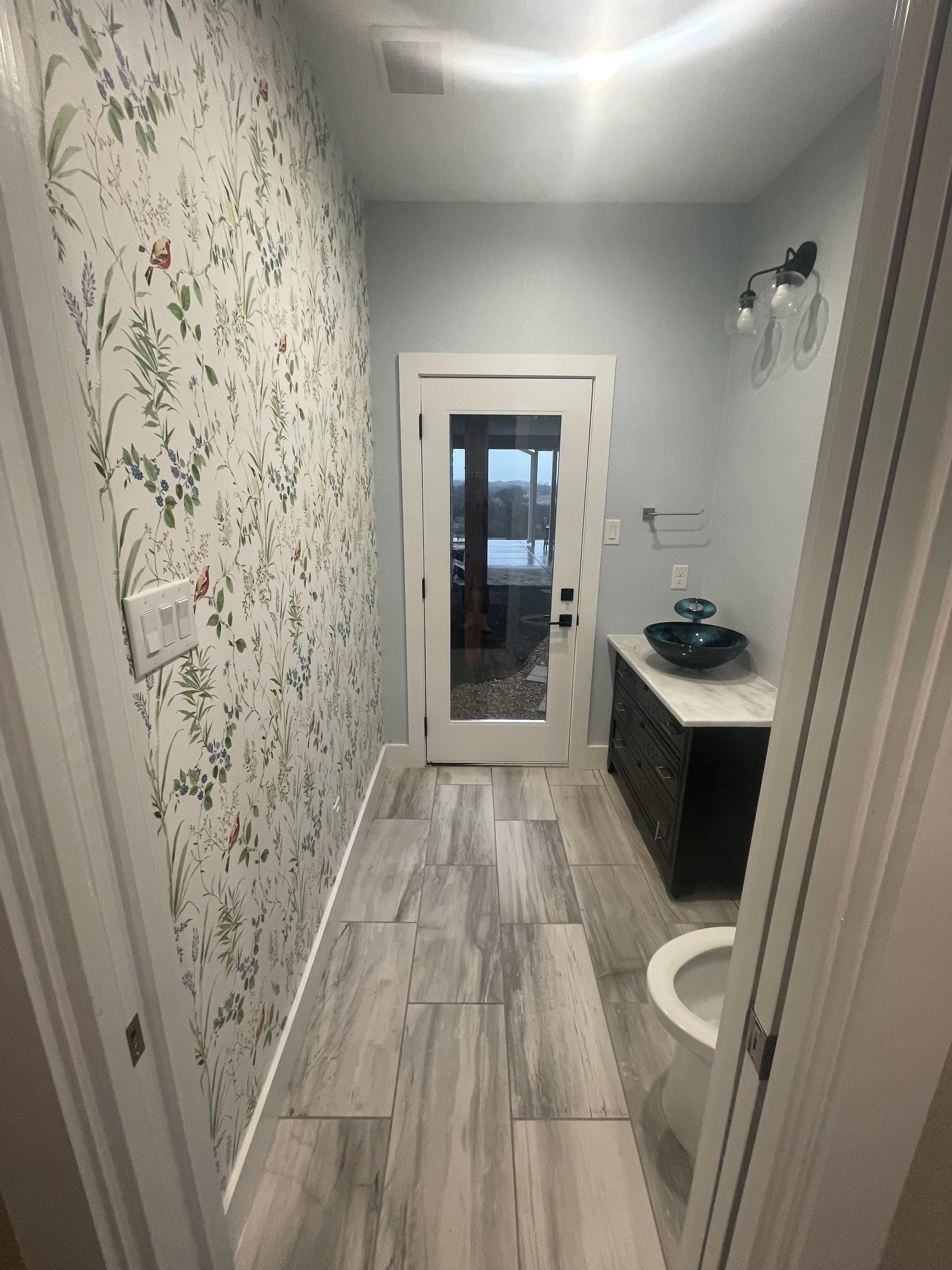A small bathroom with a toilet, a black vanity with a blue sink, and a glass door leading outside. One wall has floral wallpaper, and the other wall is painted light blue. The floor has beige and gray tiles.