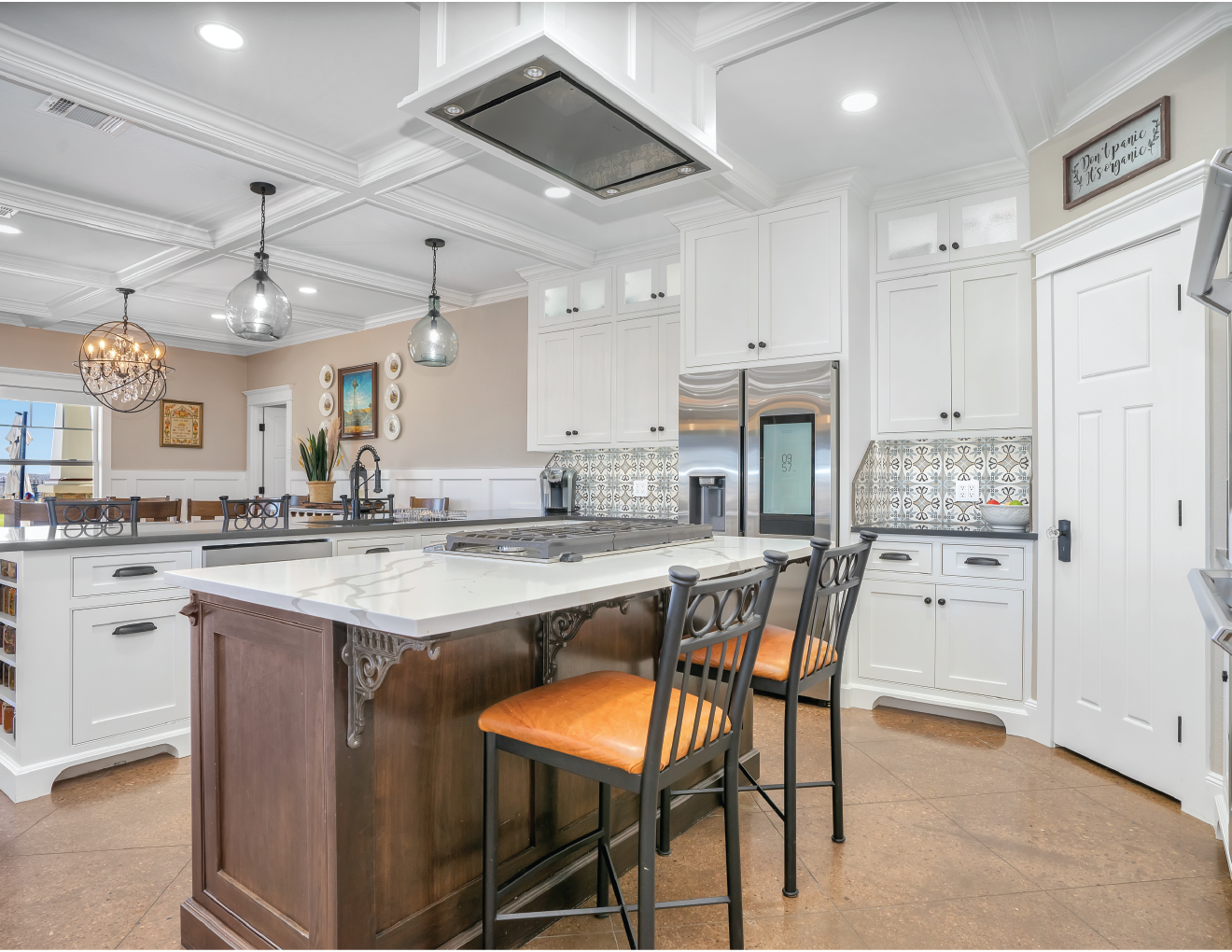 Modern kitchen with white cabinets, a central island with a dark wood base and white countertop, orange cushioned bar stools, stainless steel refrigerator, patterned backsplash, and decorative light fixtures.