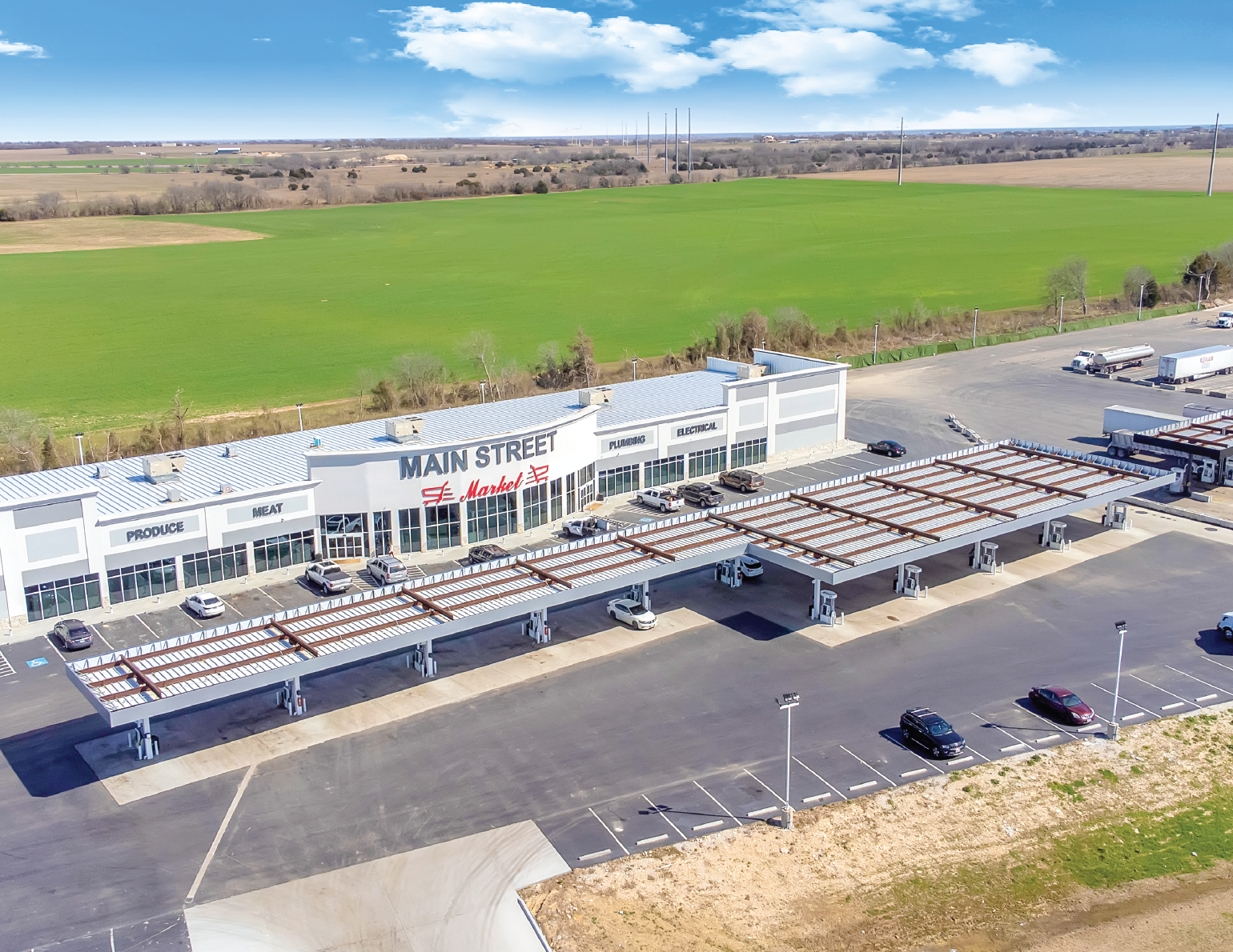 Aerial view of a modern shopping center with a parking lot. The building has a sign that reads 'Main Street Market' and sections labeled 'Produce,' 'Meat,' 'Plumbing,' and 'Electrical.' There are several cars parked in front and under a drive-thru ca