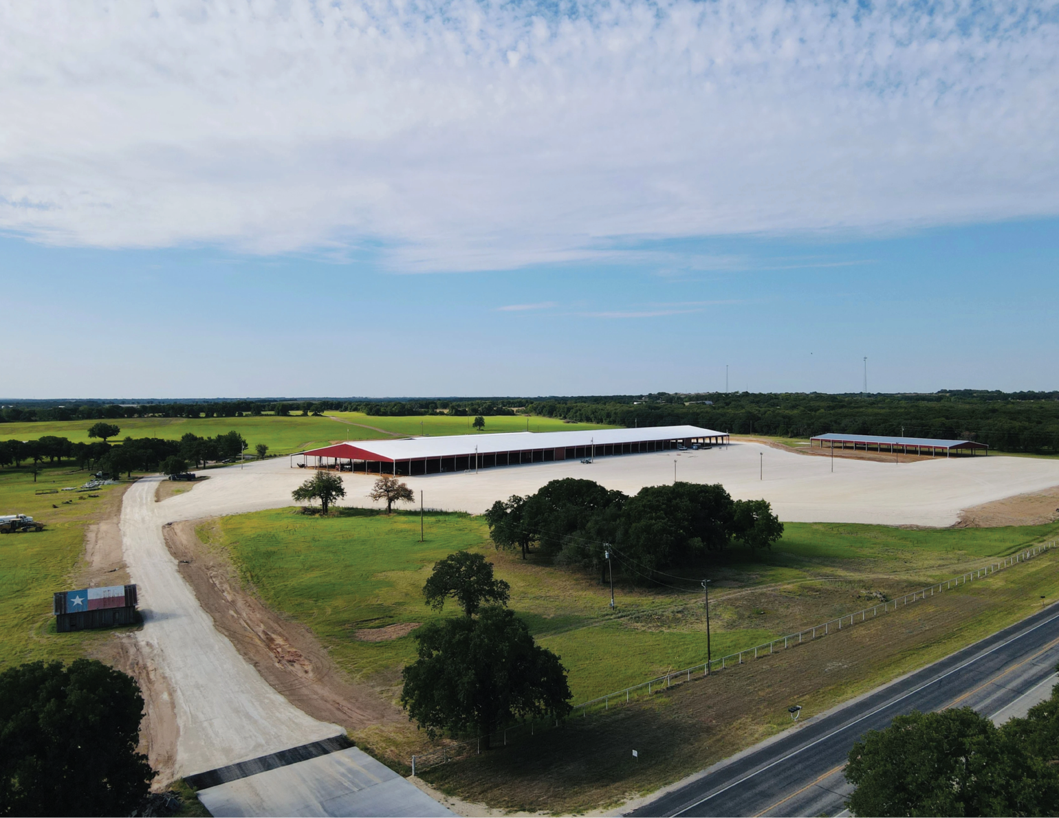 A large, open, rural area with two covered structures and a dirt path leading through green fields, surrounded by trees, and a paved road with a Texas-themed shipping container nearby.