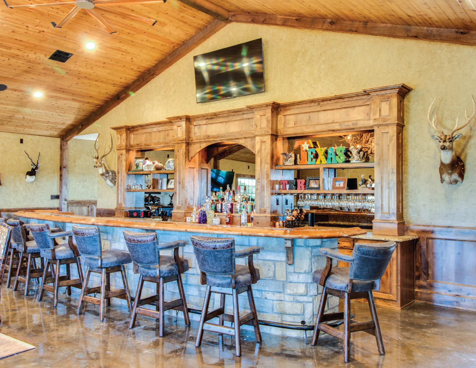 Interior of a rustic bar with a wooden counter and high chairs, decorated with mounted deer heads and holiday decorations, with a TV mounted on the wall.
