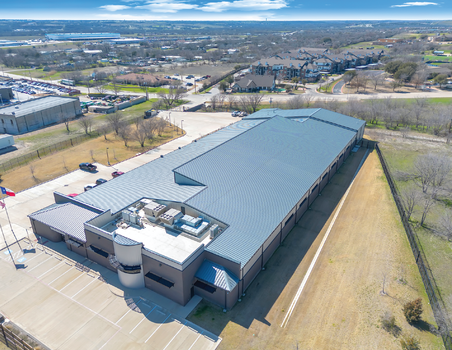 An aerial view of a large commercial building with a light blue metal roof, parking lot with some cars, surrounded by a fenced area with grass and trees, in a suburban setting with other residential and industrial buildings nearby.
