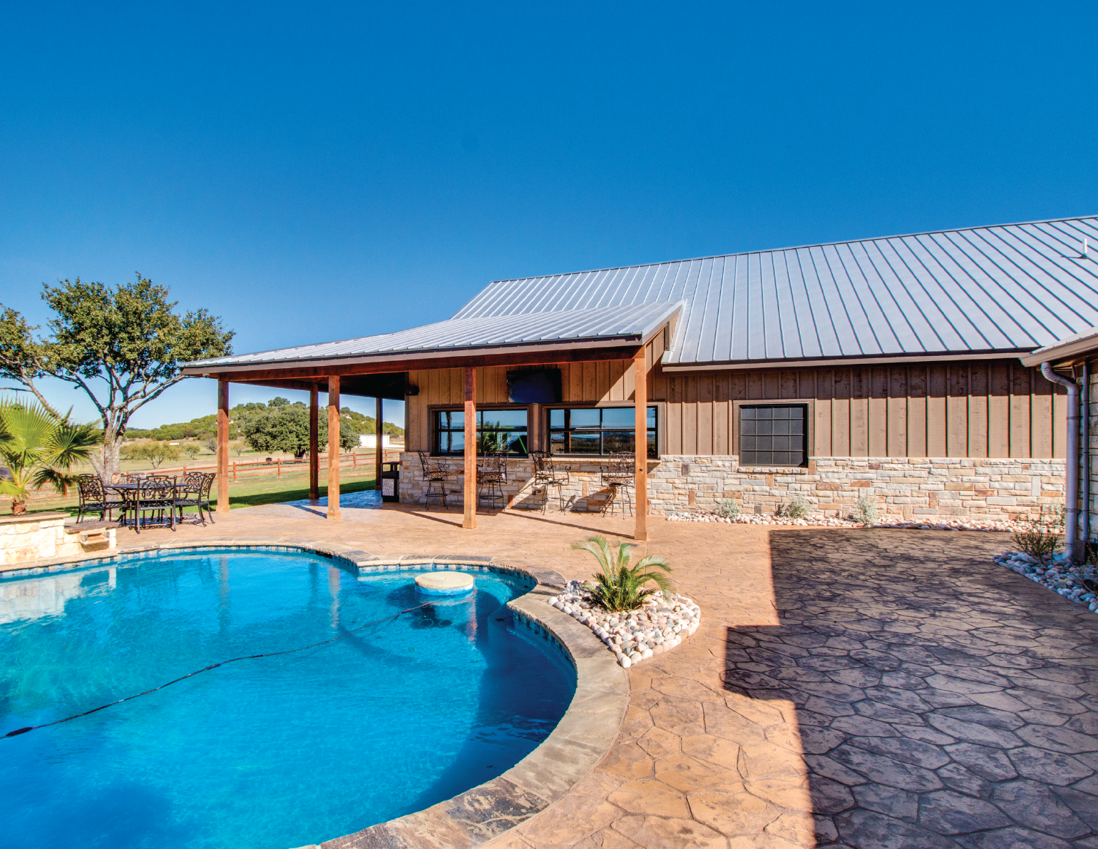 A backyard with a swimming pool, stone-paved patio, a house with a wooden exterior and a metal roof, surrounded by trees and open land under a clear blue sky.