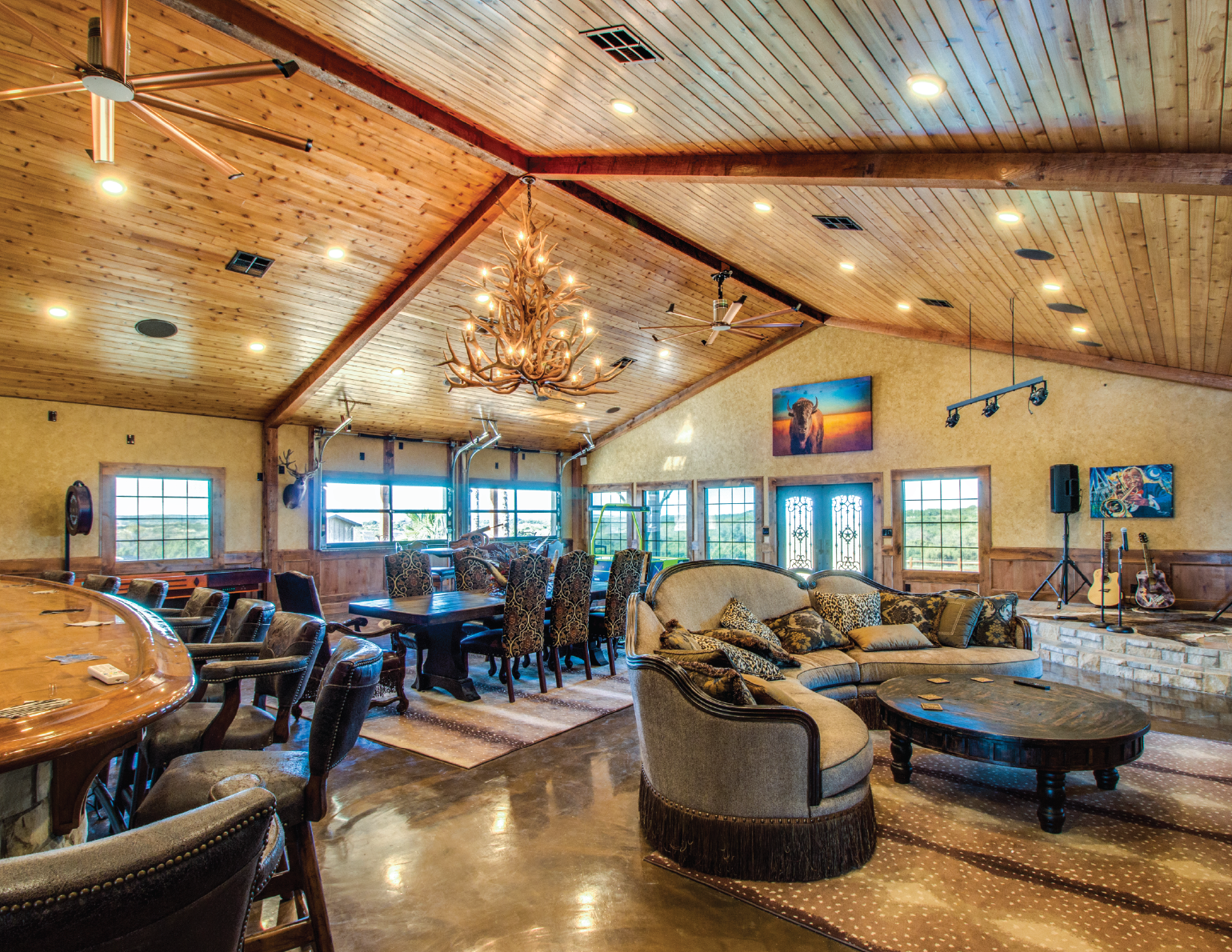 A spacious indoor lounge with a wooden ceiling and walls, furnished with a large sofa, armchairs, a dining table with chairs, and a decorated room with wall art, guitars, and a stage area.