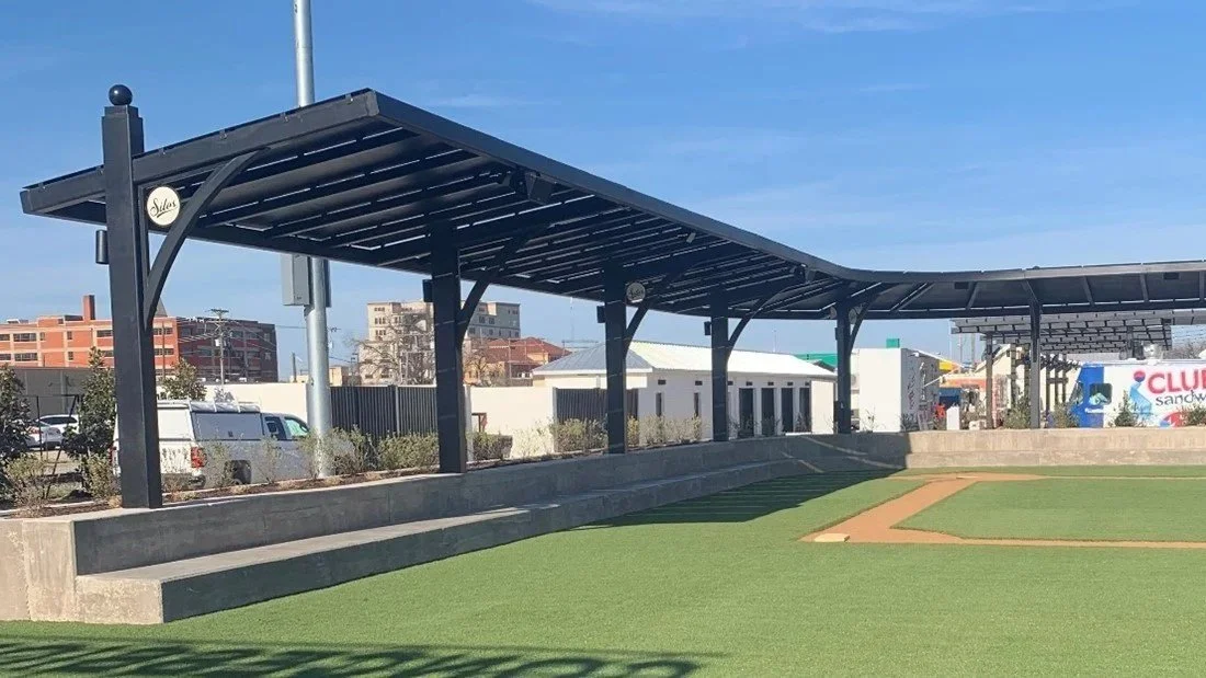 Empty baseball field dugout with a metal roof, artificial turf, and city buildings in the background.