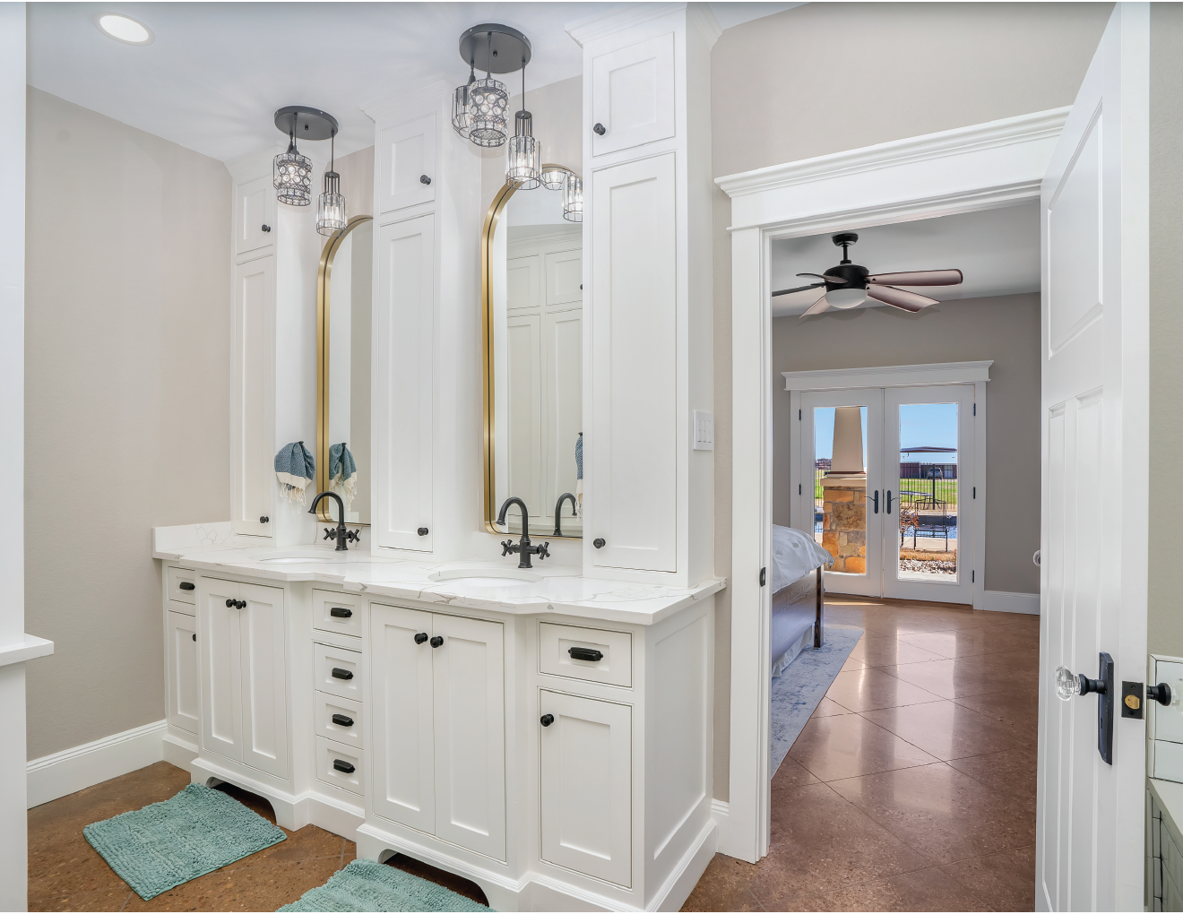 Bathroom with white vanity, three mirrors with gold frames, black faucets, pendant lights, and blue towels, with a view into a bedroom with a ceiling fan and sliding glass doors leading outside.