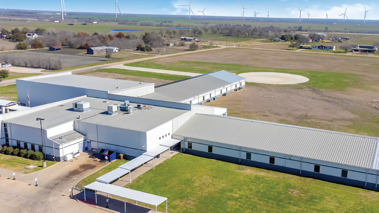 Aerial view of a large industrial or educational campus with white buildings and metal roofs, surrounded by fields and roads. Wind turbines are visible in the background.