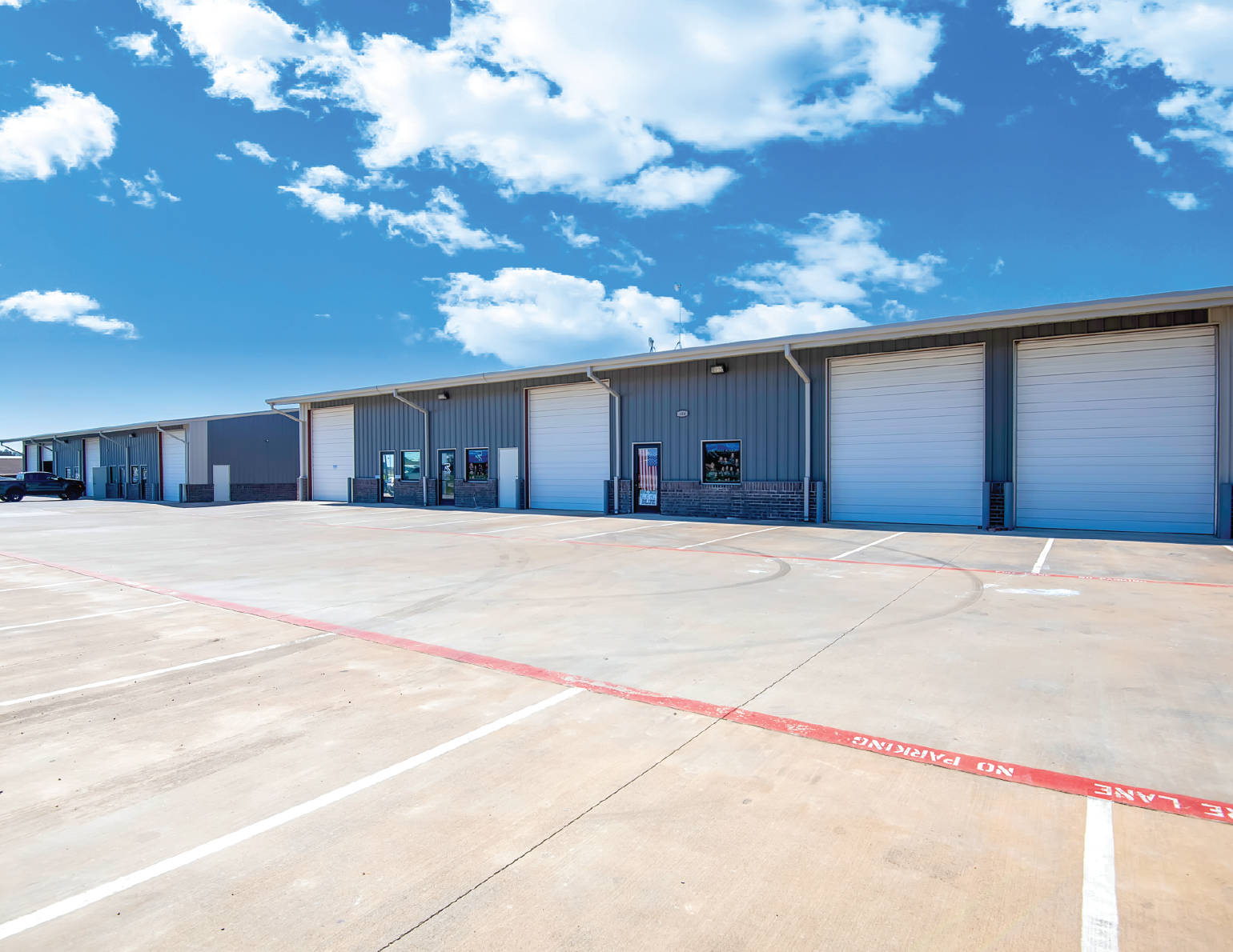 Empty parking lot in front of a large industrial warehouse with multiple roll-up doors under a partly cloudy sky.
