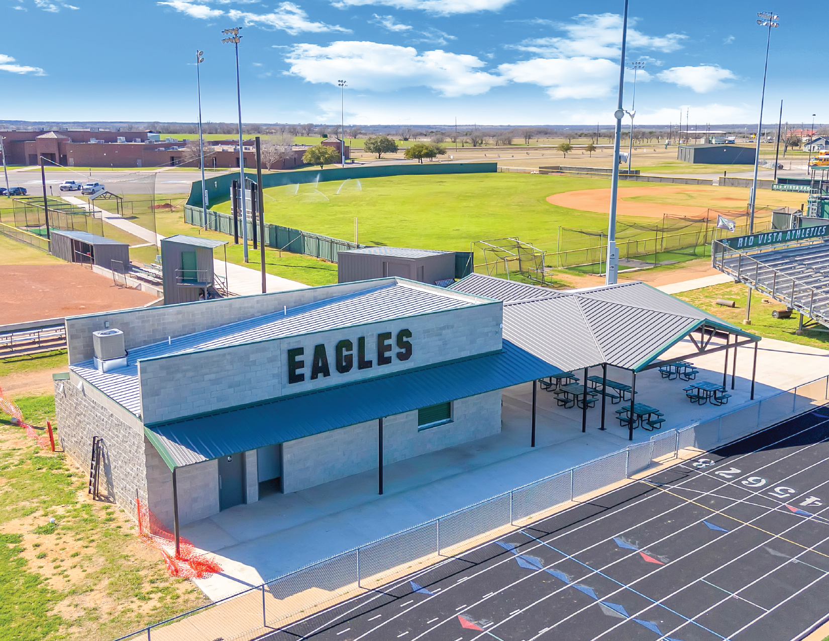 Aerial view of a sports complex with a building labeled 'EAGLES', a baseball field, and a parking lot, under a partly cloudy blue sky.