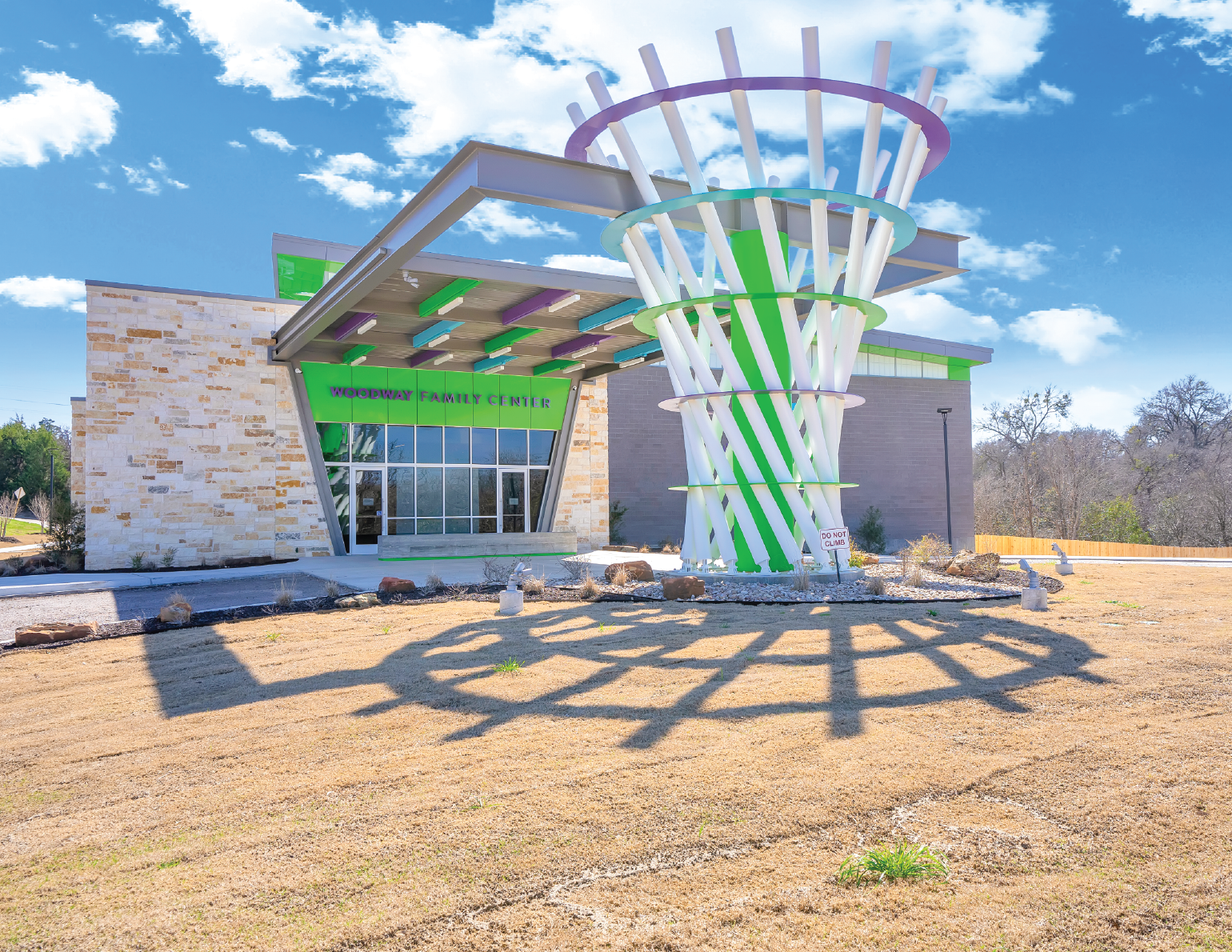 The Woodway Family Center building features a modern design with an angular entrance, stone facade, and large glass windows. In front, there is a large white and green playful sculpture casting shadows on the dry grass, with a sign reading 'Do Not Cl