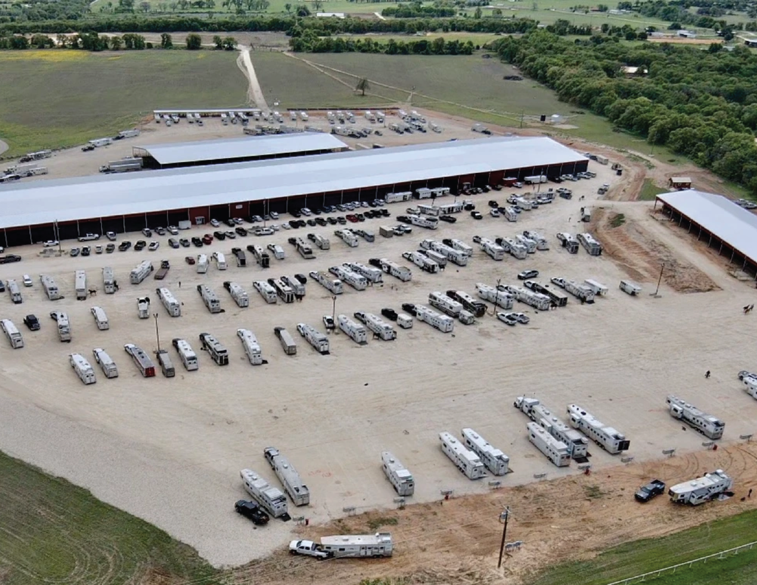 An aerial view of a large cluster of RVs and vehicles parked on a dirt lot near a long red and white building, with trees and fields surrounding the area.