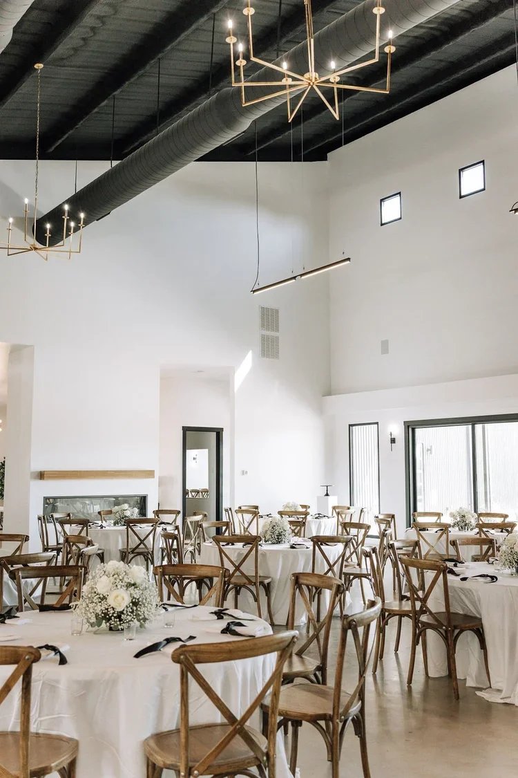 A wedding or event reception hall with round tables covered in white tablecloths, decorated with floral centerpieces, surrounded by wooden chairs, and modern lighting fixtures hanging from a black ceiling.