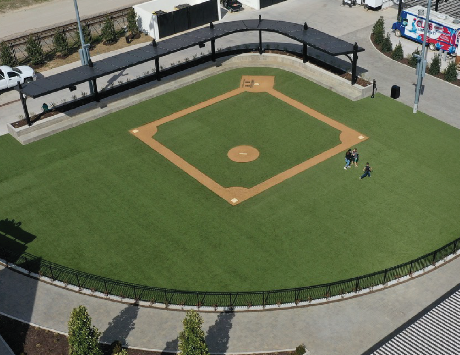 A baseball field with a grassy outfield and a dirt infield, surrounded by a black metal fence and a concrete walkway, with three children running on the grass.