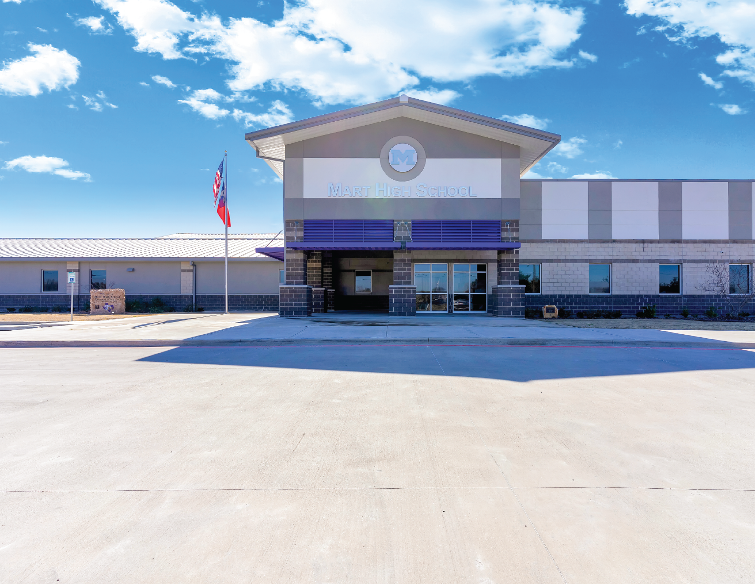 Front view of Mart High School building with a flagpole flying the American flag, under a partly cloudy sky.