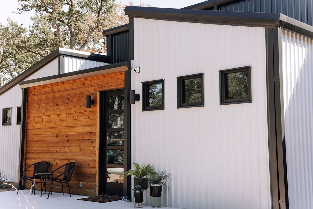 Modern house with white and wood exterior, three small square windows, and a small porch with two chairs and potted plants.