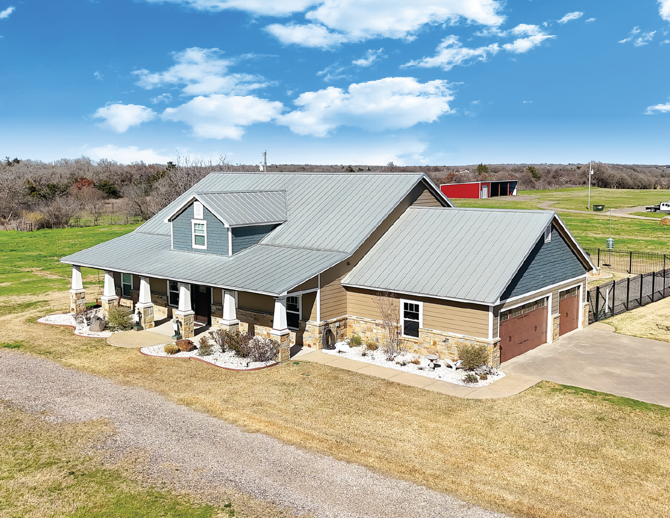 A large house with a metal roof, front porch, and attached two-car garage, surrounded by a lawn and open fields under a blue sky with clouds.